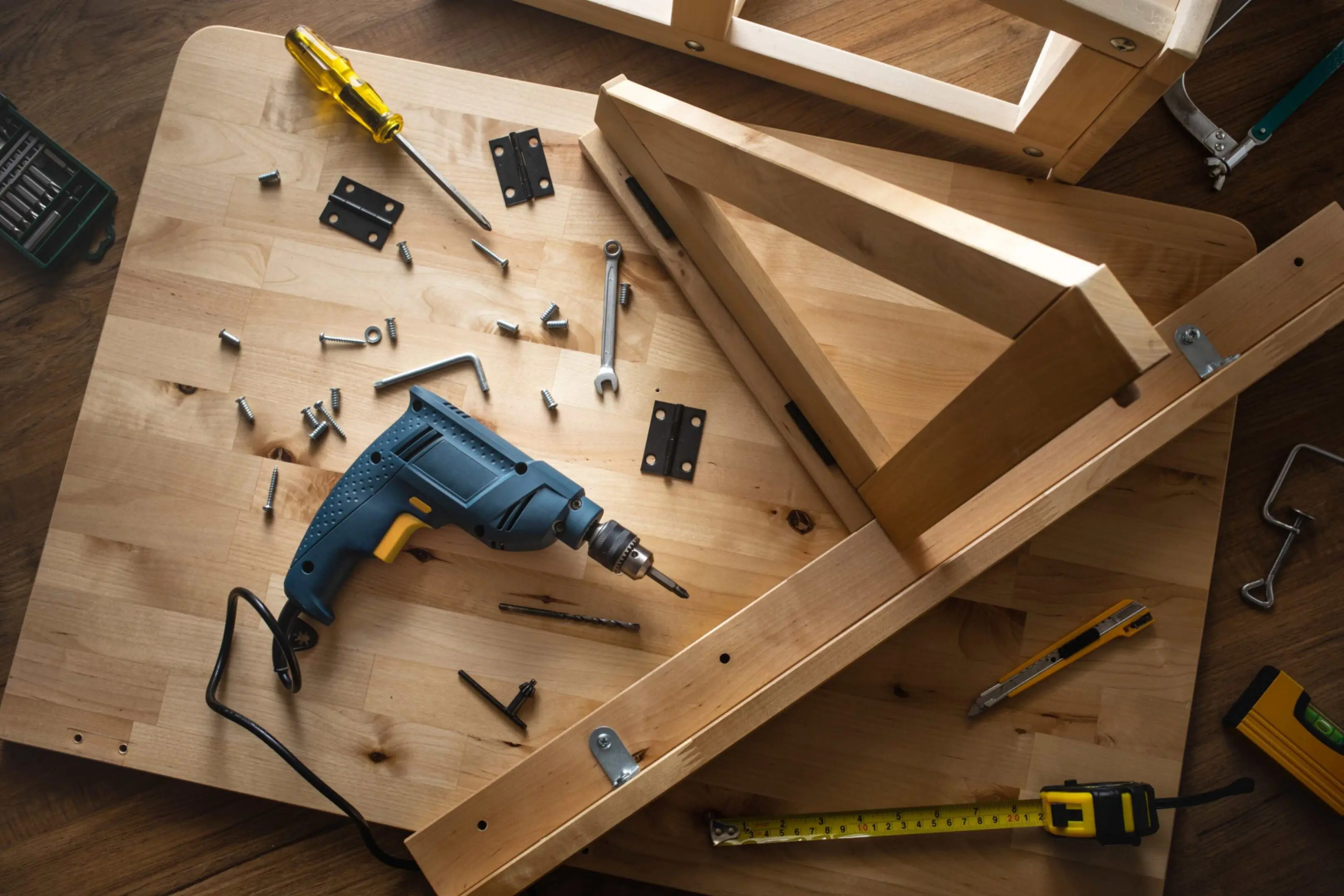 jpeg-optimizer_GettyImages-1067496776 Wooden furniture pieces with a drill, screws, screwdriver, wrench, utility knife, and measuring tape on a wooden surface.